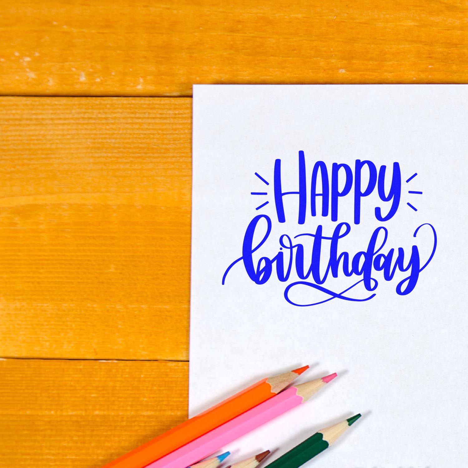 Happy Birthday rubber stamp design on white card with colored pencils on a wooden table, ideal for birthday cards and crafts.