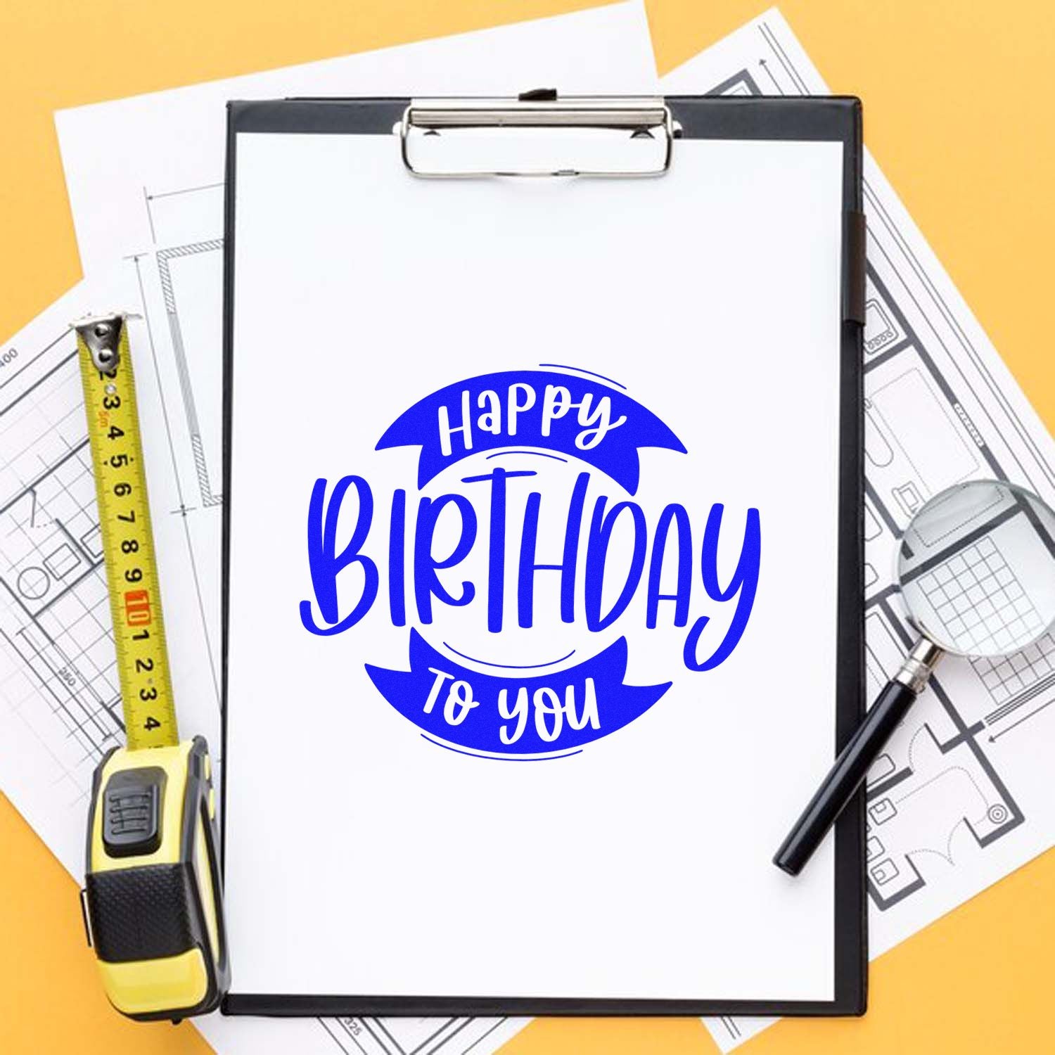 Custom Happy Birthday rubber stamp mockup with clipboard, tape measure, and floor plan drawings for business or craft use.