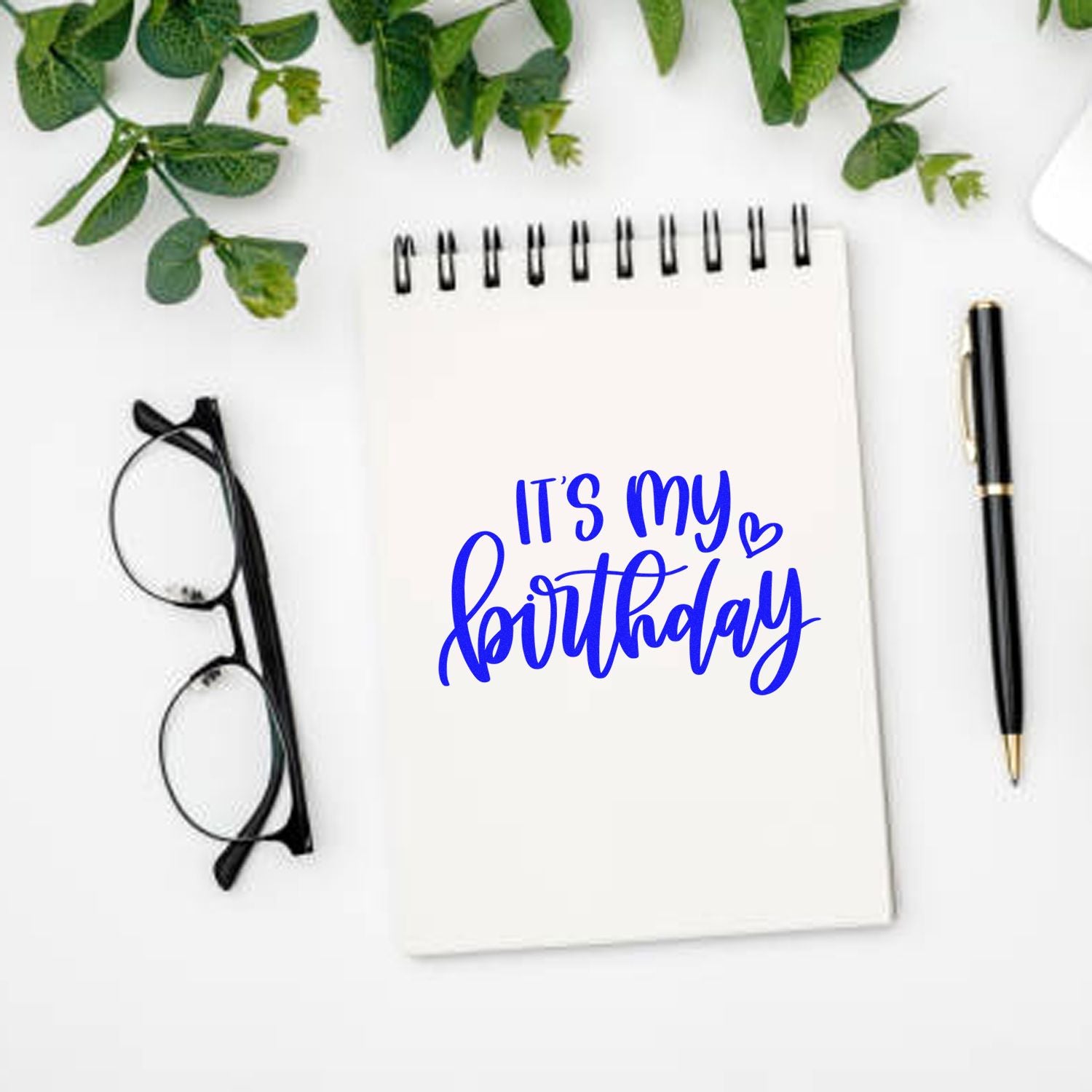 Its My Birthday rubber stamp design on a notebook, styled with pen, glasses and greenery for a cheerful birthday craft display.