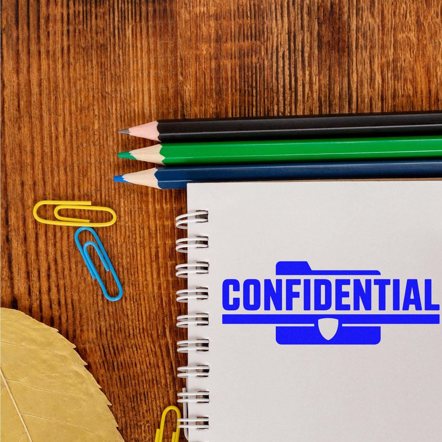 Confidential rubber stamp on notebook page, with pencils and paper clips, for office document security and file marking.