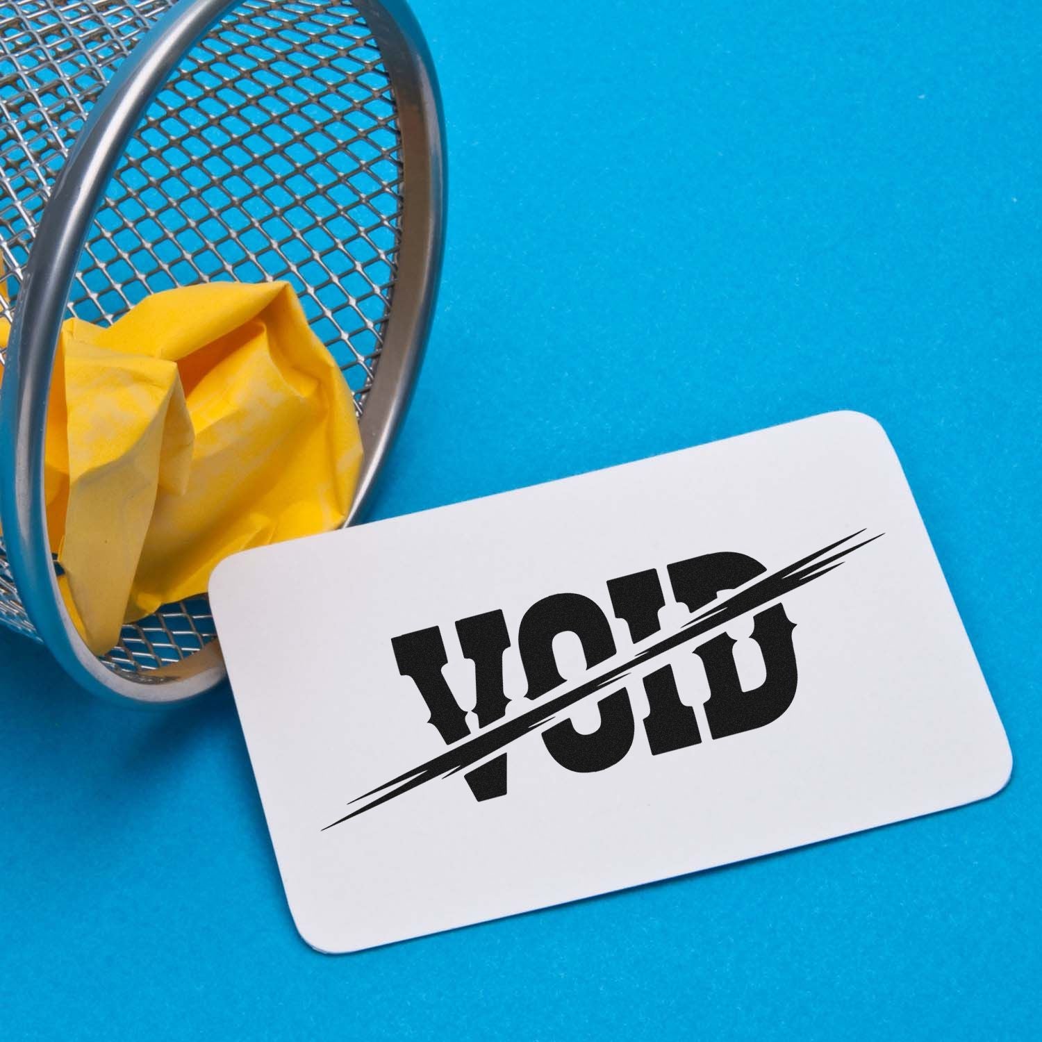 VOID rubber stamp impression on a white card beside a waste basket, showing document cancellation and rejection.