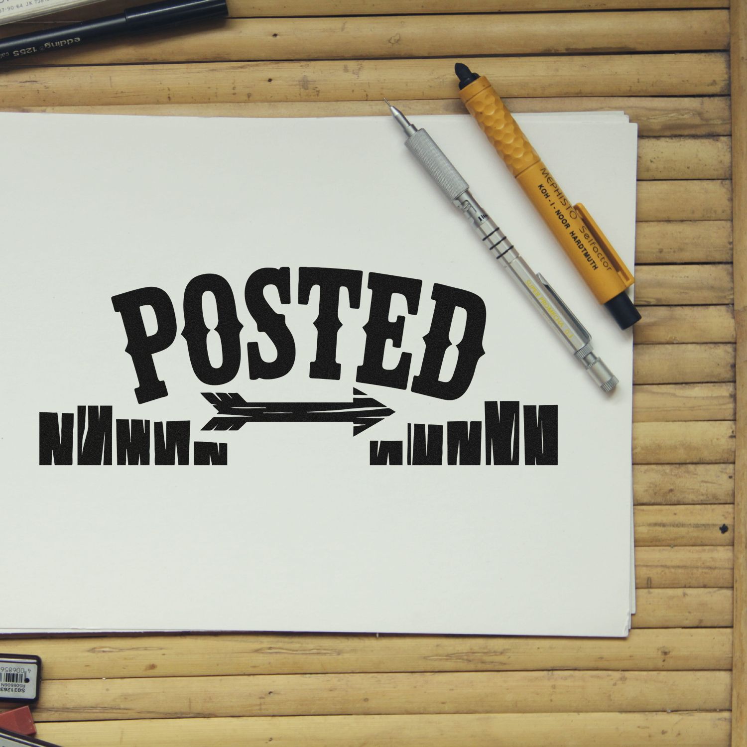 Custom rubber stamp design with bold POSTED text and arrow on paper, beside drafting tools on a wooden desk.