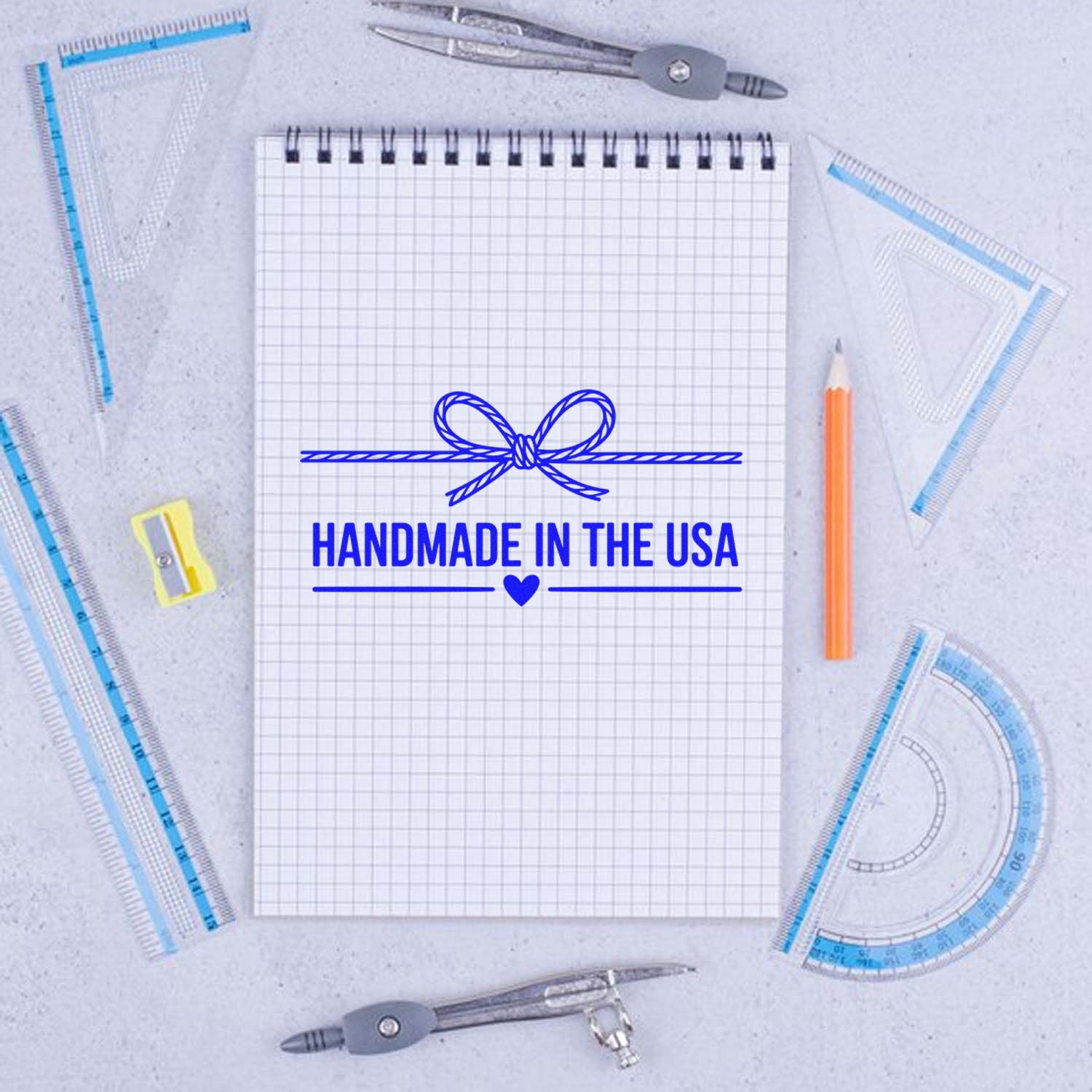 Handmade in the USA rubber stamp design on grid notepad, surrounded by drafting tools and pencils for craft branding.