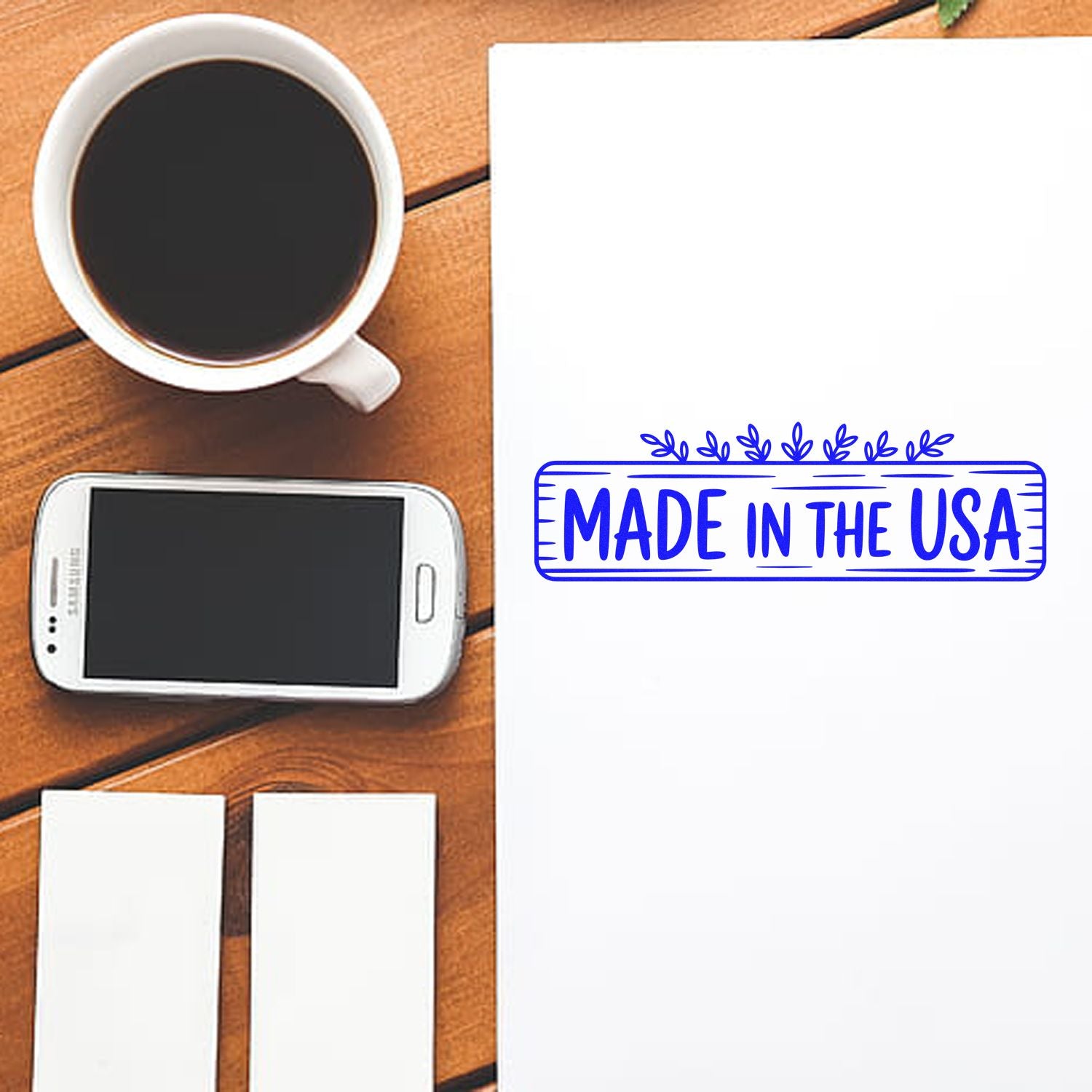 Made in the USA rubber stamp impression on white paper beside coffee, smartphone, and blank labels on a wooden desk.