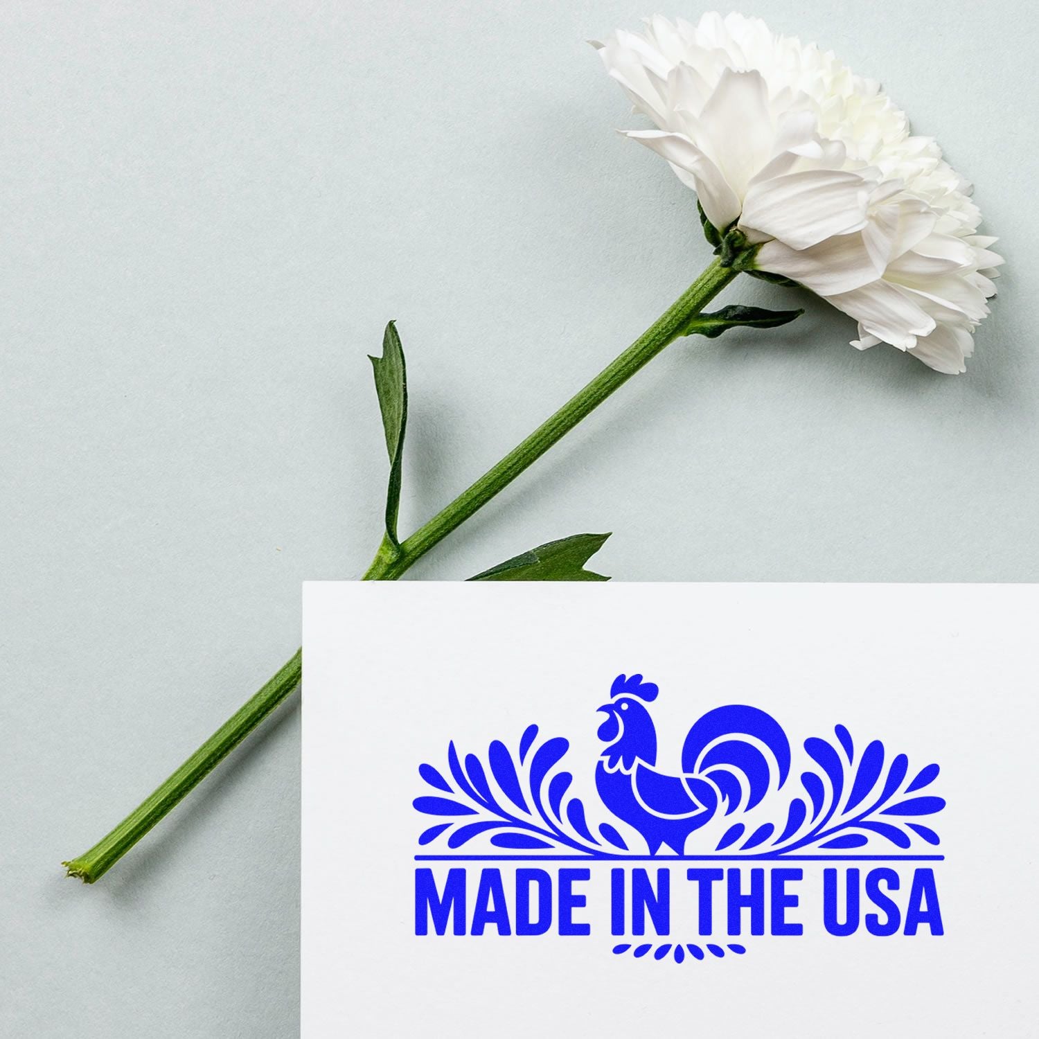Made in the USA rubber stamp featuring a blue rooster design on white card, styled with a white flower backdrop.