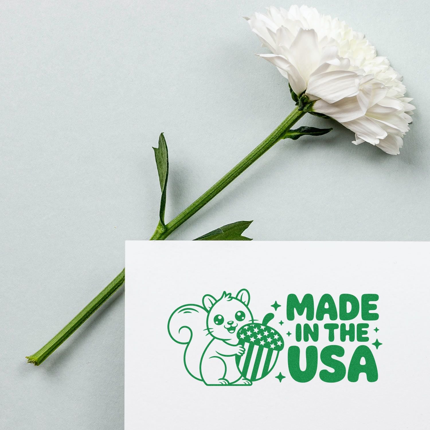 Green Made in the USA rubber stamp with squirrel and acorn on a white card, shown beside a white flower.