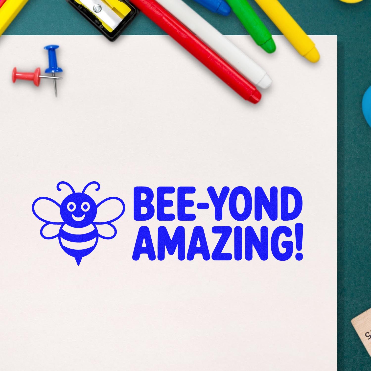 Blue bee rubber stamp reading Bee-Yond Amazing! on paper, surrounded by markers and push pins for craft or teacher use.