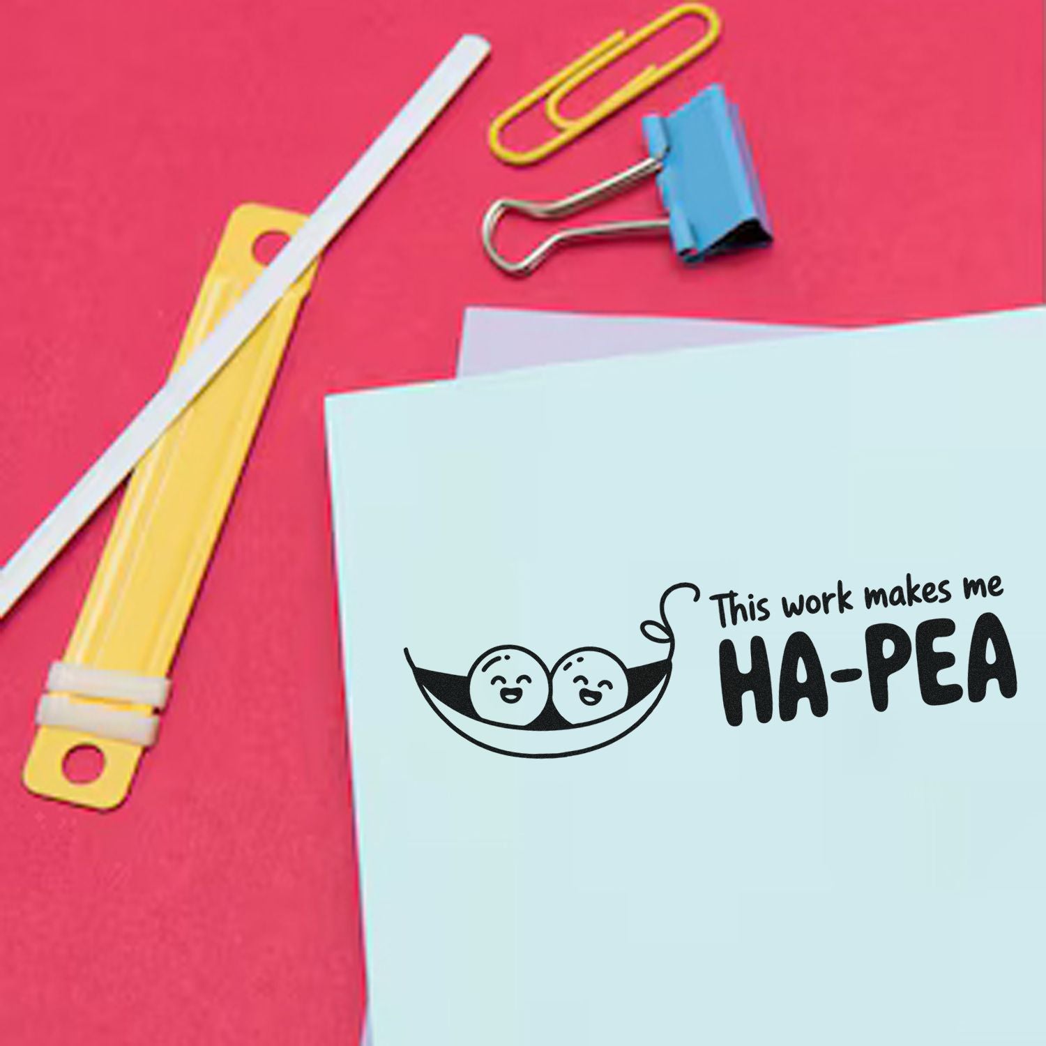 Humorous rubber stamp design on paper reading This work makes me HA-PEA with paper clips and stationery on a pink desk.