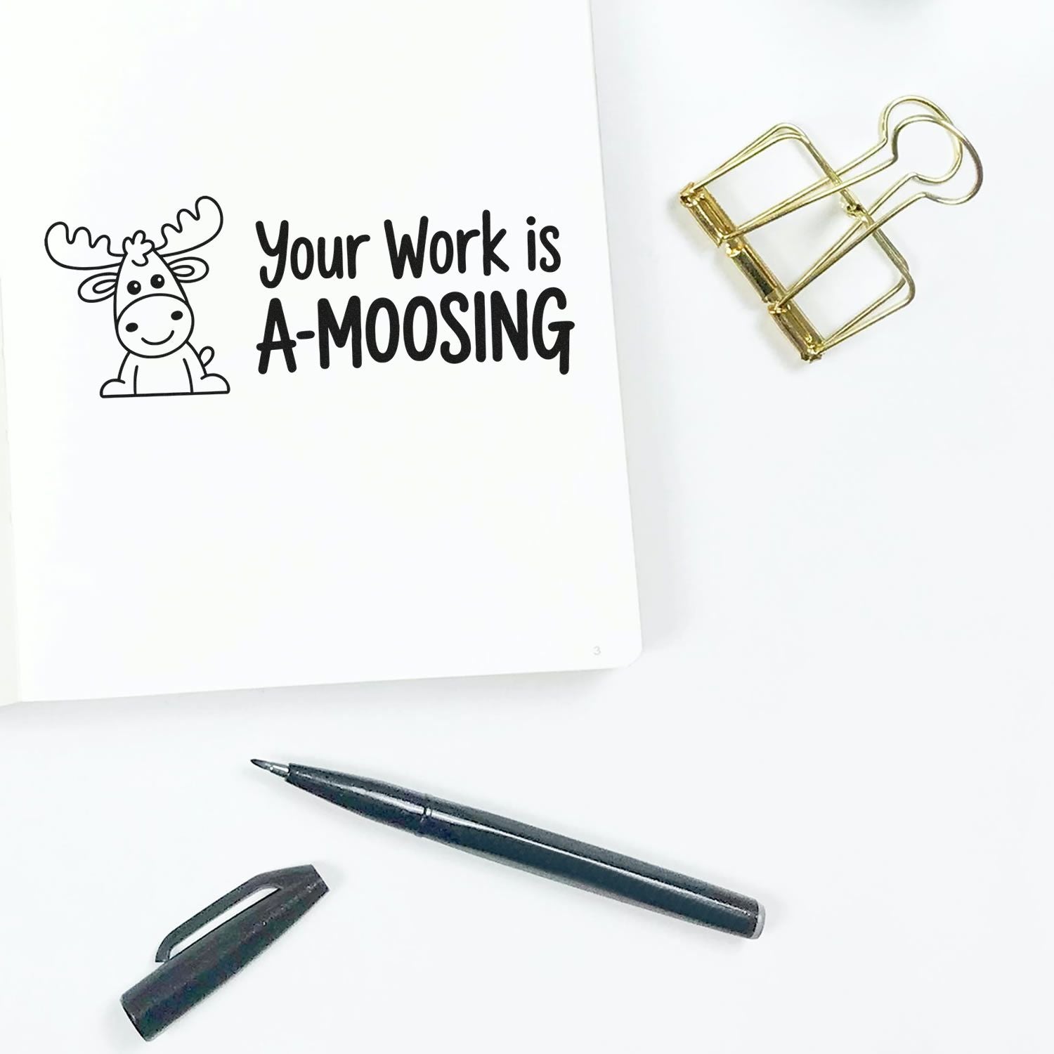 Moose-themed rubber stamp impression reading Your Work is A-MOOSING on notebook with pen and paper clip.