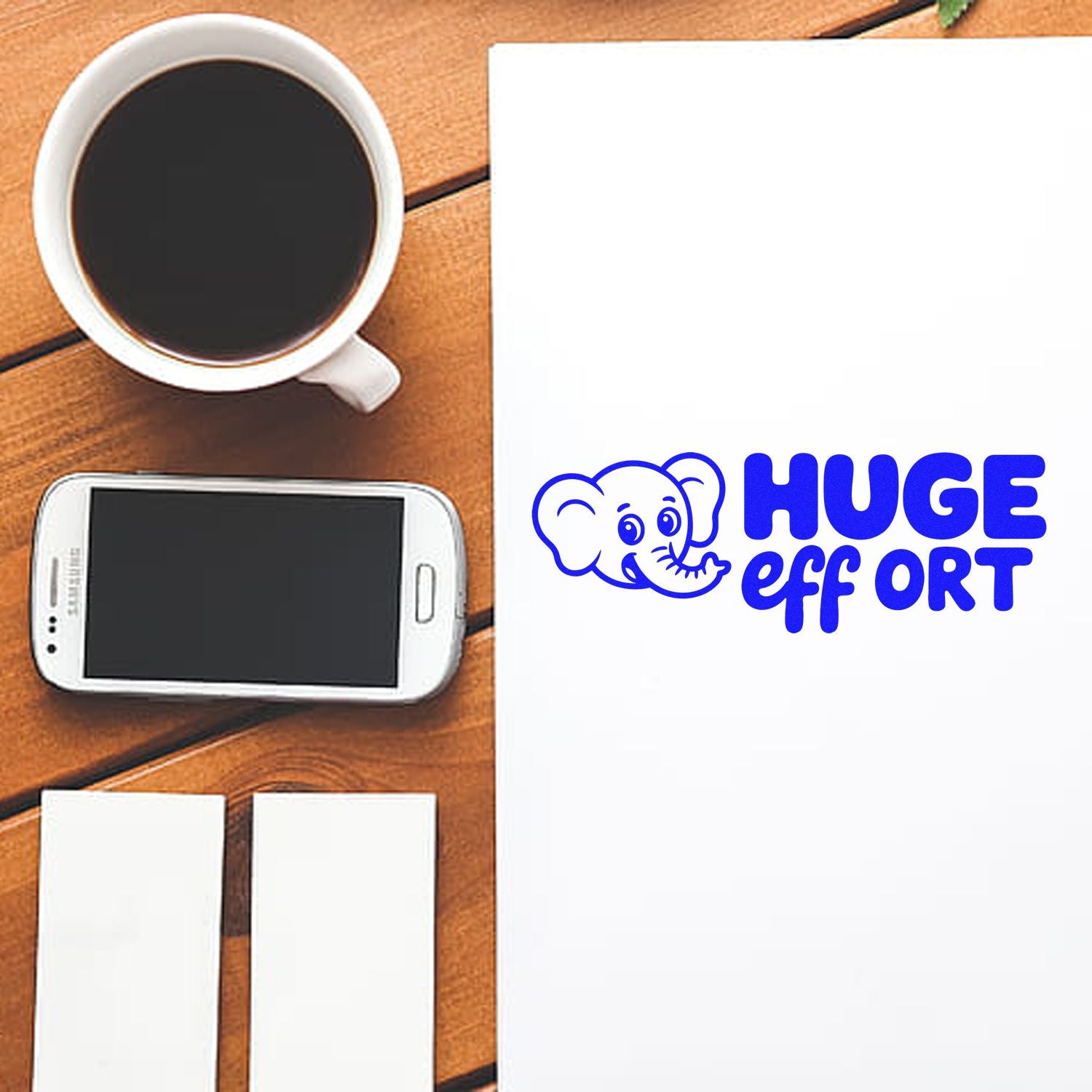 Custom rubber stamp mockup with blue elephant and Huge Effort text on a blank sheet beside coffee and phone.