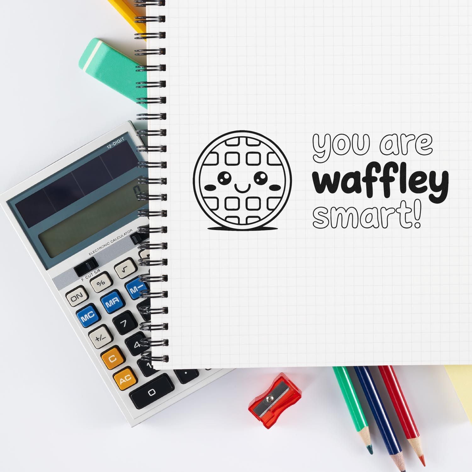 Rubber stamp design with a smiling waffle and you are waffley smart! on a notebook, styled with school supplies and calculator.