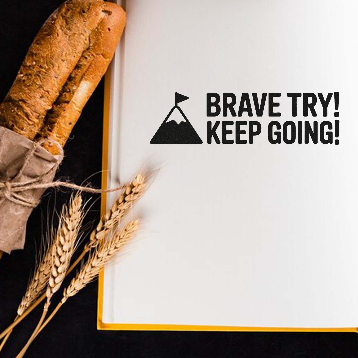 Motivational rubber stamp design with mountain icon and Brave Try! Keep Going! text on a notebook beside bread and wheat.