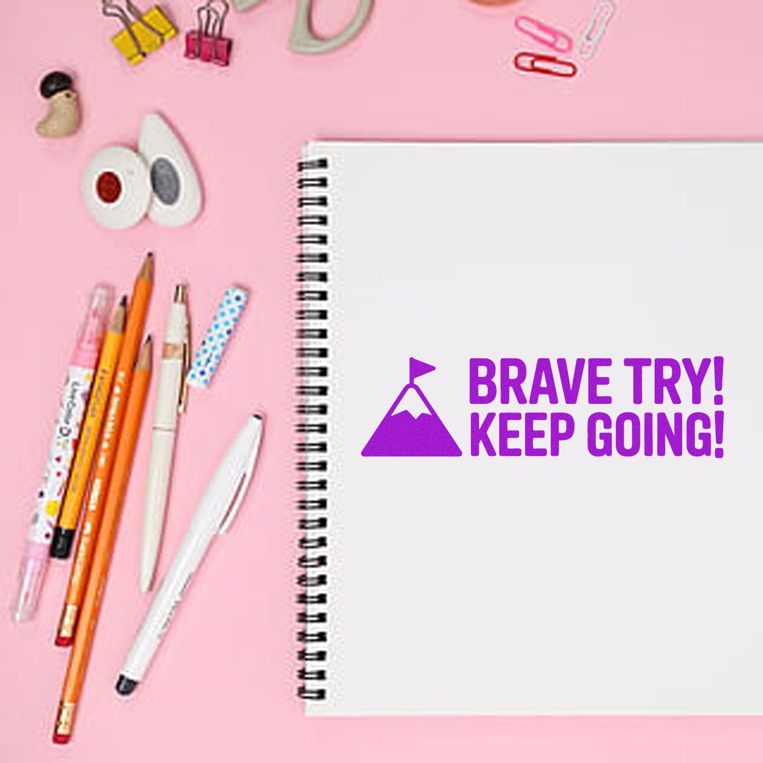 Motivational rubber stamp on spiral notebook reading Brave Try! Keep Going! with stationery on pink desk.
