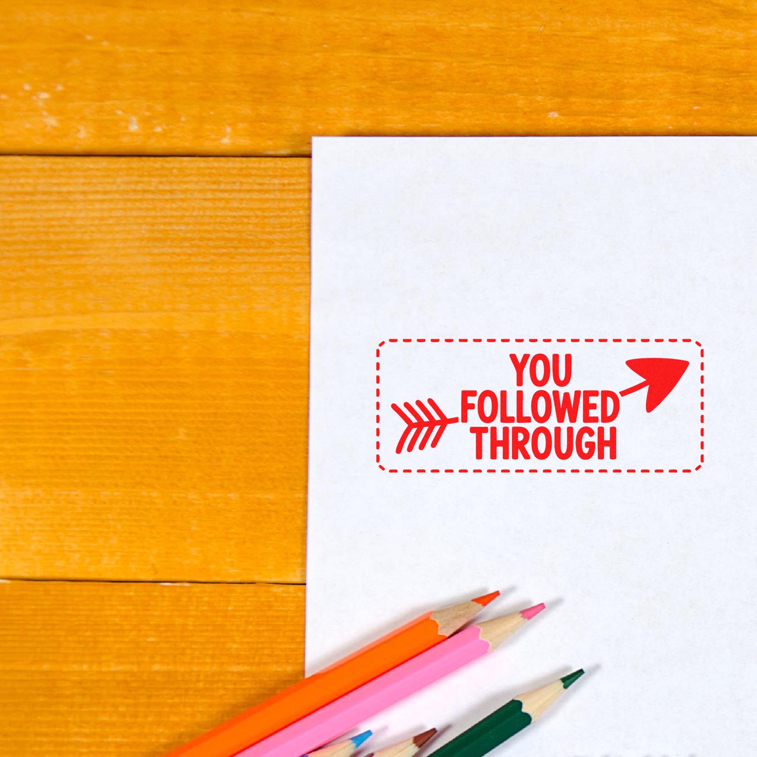 Custom rubber stamp reading You Followed Through on white paper with colored pencils on a wooden desk.