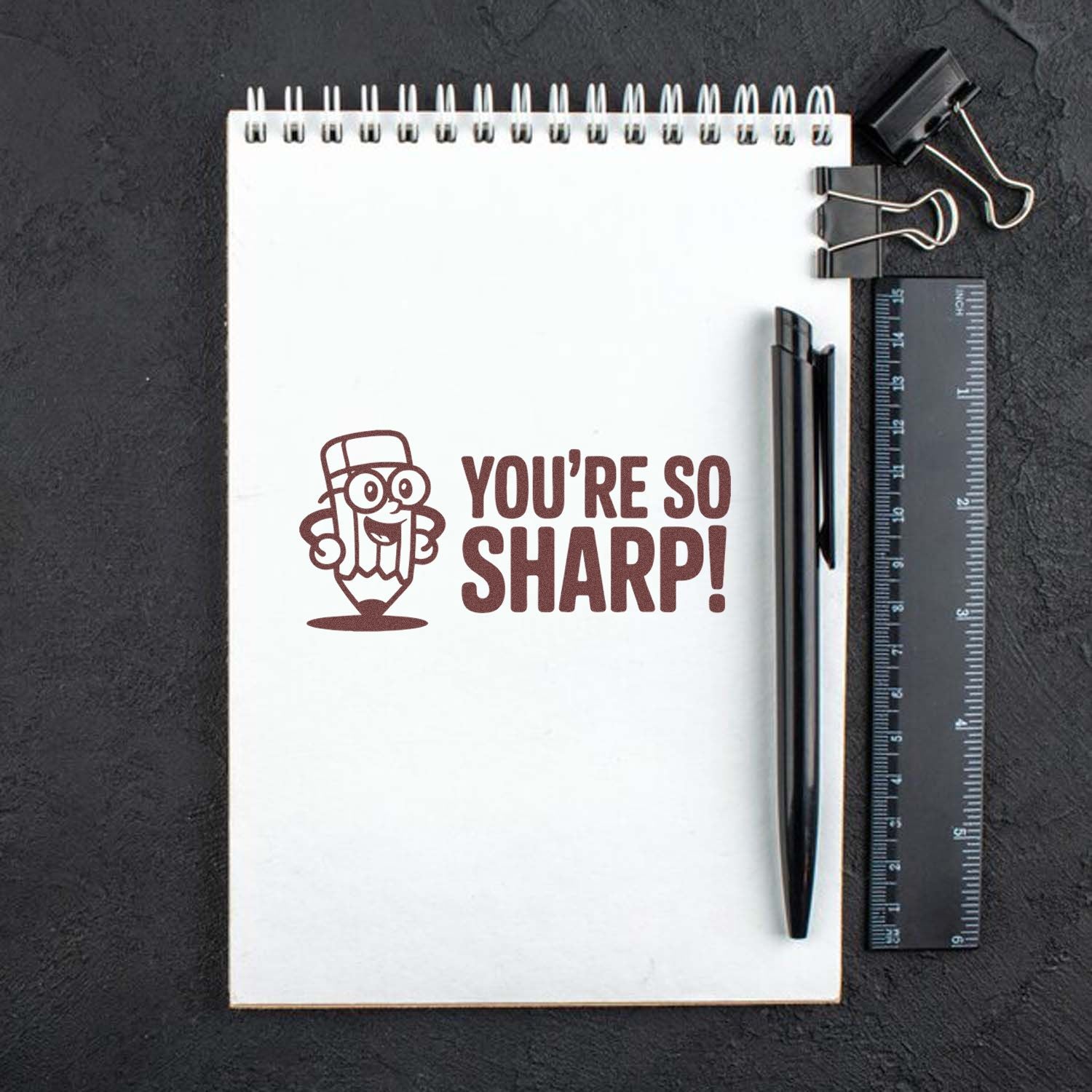 "You're So Sharp!" pencil rubber stamp on a notebook with pen and ruler, ideal for teacher rewards and stationery branding.