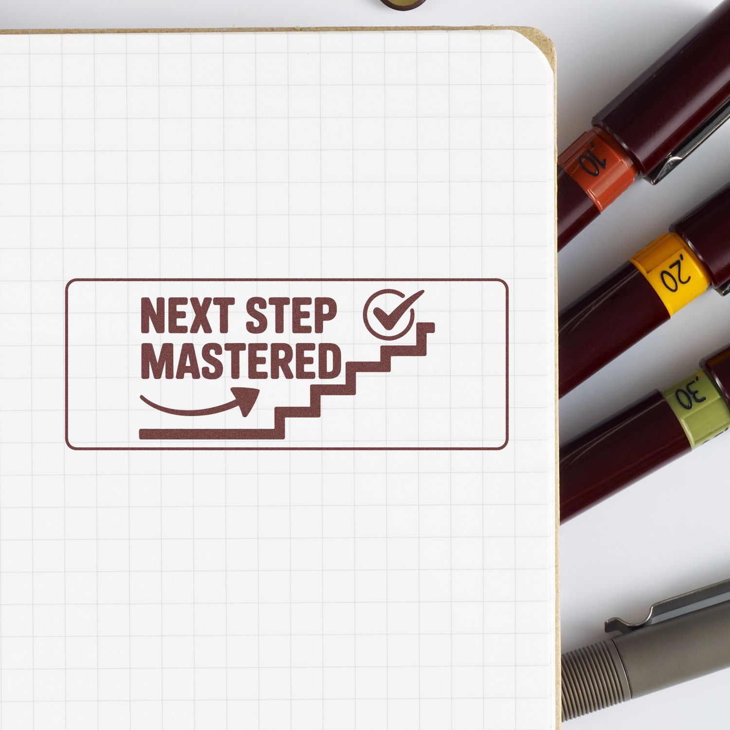 Motivational rubber stamp reading Next Step Mastered with staircase checkmark on notebook, shown with color-coded markers.