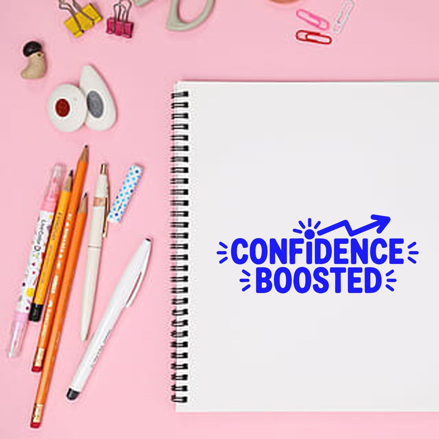 Custom rubber stamp design reading Confidence Boosted on a spiral notebook with stationery on a pink background.