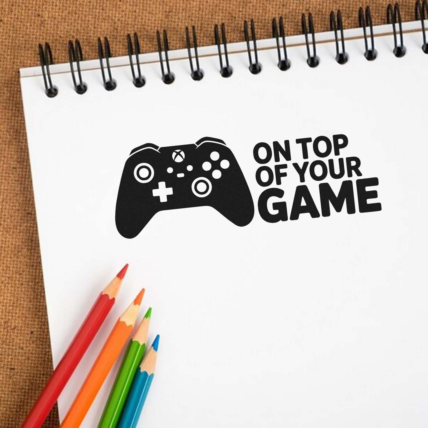 Gaming rubber stamp design with controller icon and On Top of Your Game slogan on a notebook mockup.