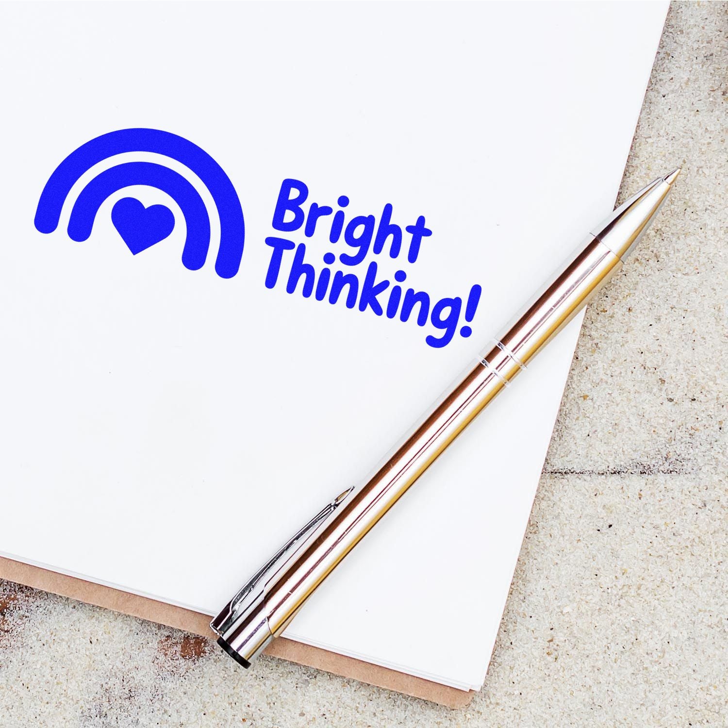 Blue Bright Thinking rubber stamp with rainbow heart logo on notebook beside pen