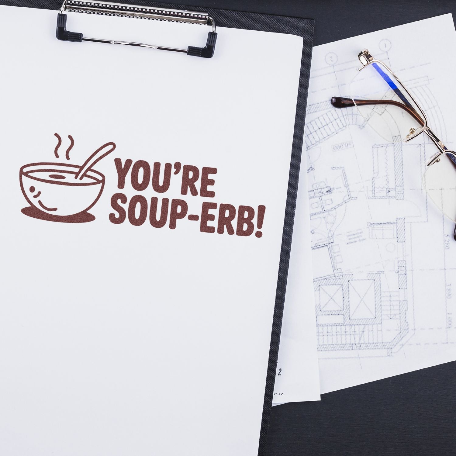 Custom rubber stamp impression reading Youre soup-erb! with soup bowl icon on paper beside eyeglasses and design sketches.