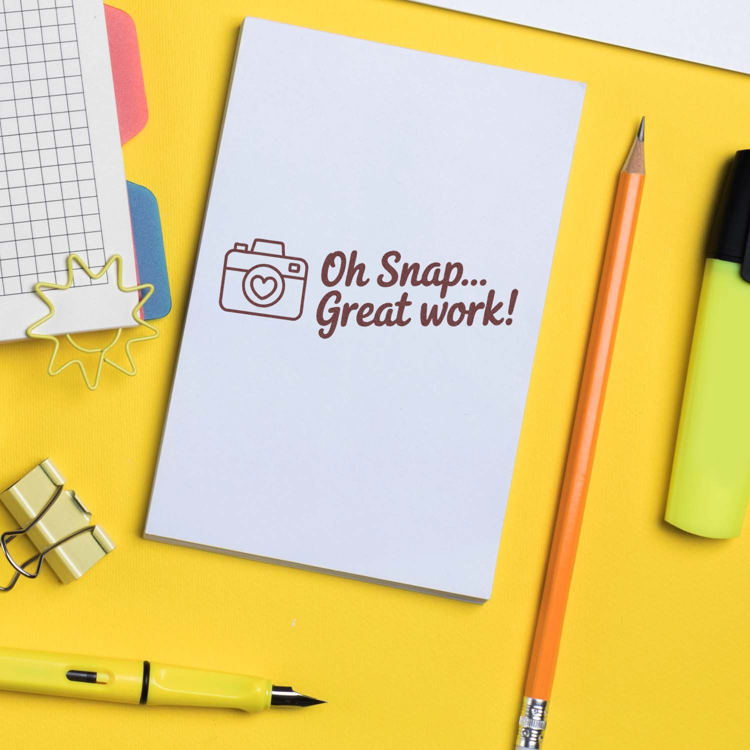 Custom rubber stamp impression reading Oh Snap... Great work! on a notebook, with camera icon on a bright yellow desk.