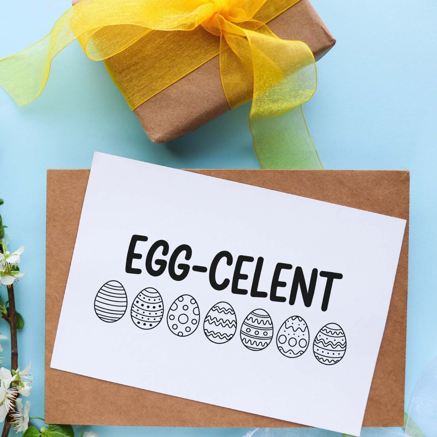 Easter rubber stamp design reading Egg-cellent with decorated eggs on a gift tag beside a wrapped present.
