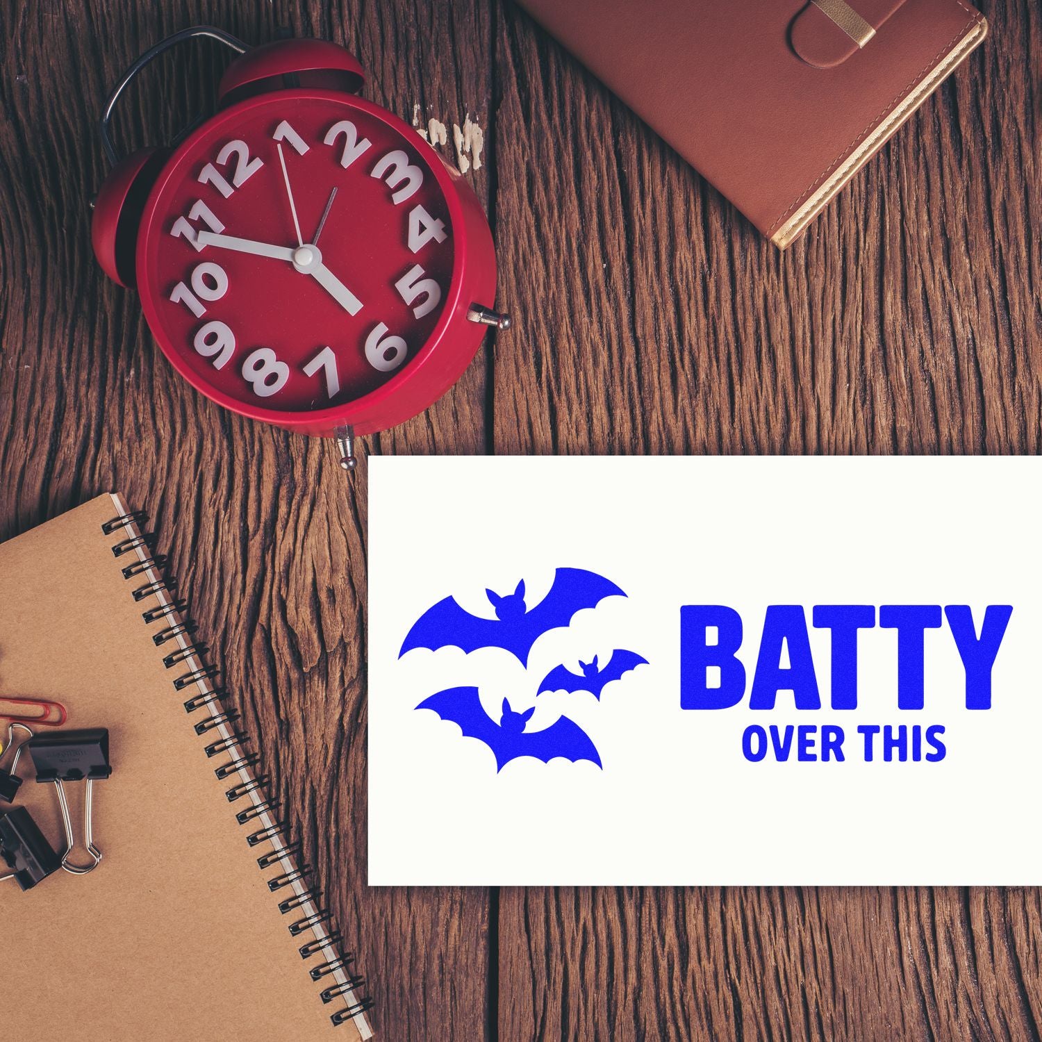 Bat-themed custom rubber stamp design with Batty Over This text, shown on a desk with notebooks and a clock.