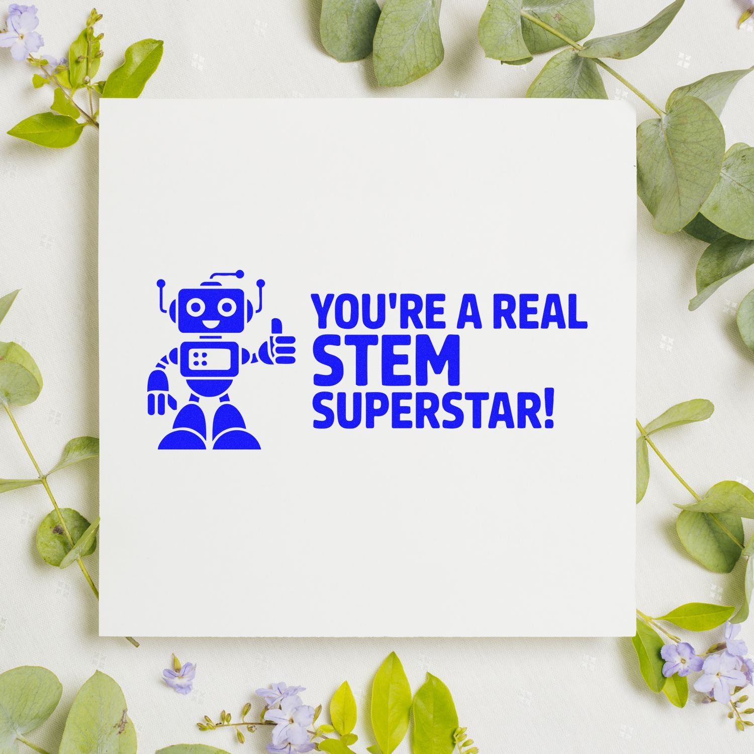 Blue robot rubber stamp design reading Youre a real STEM superstar! on a white card with greenery and flowers.