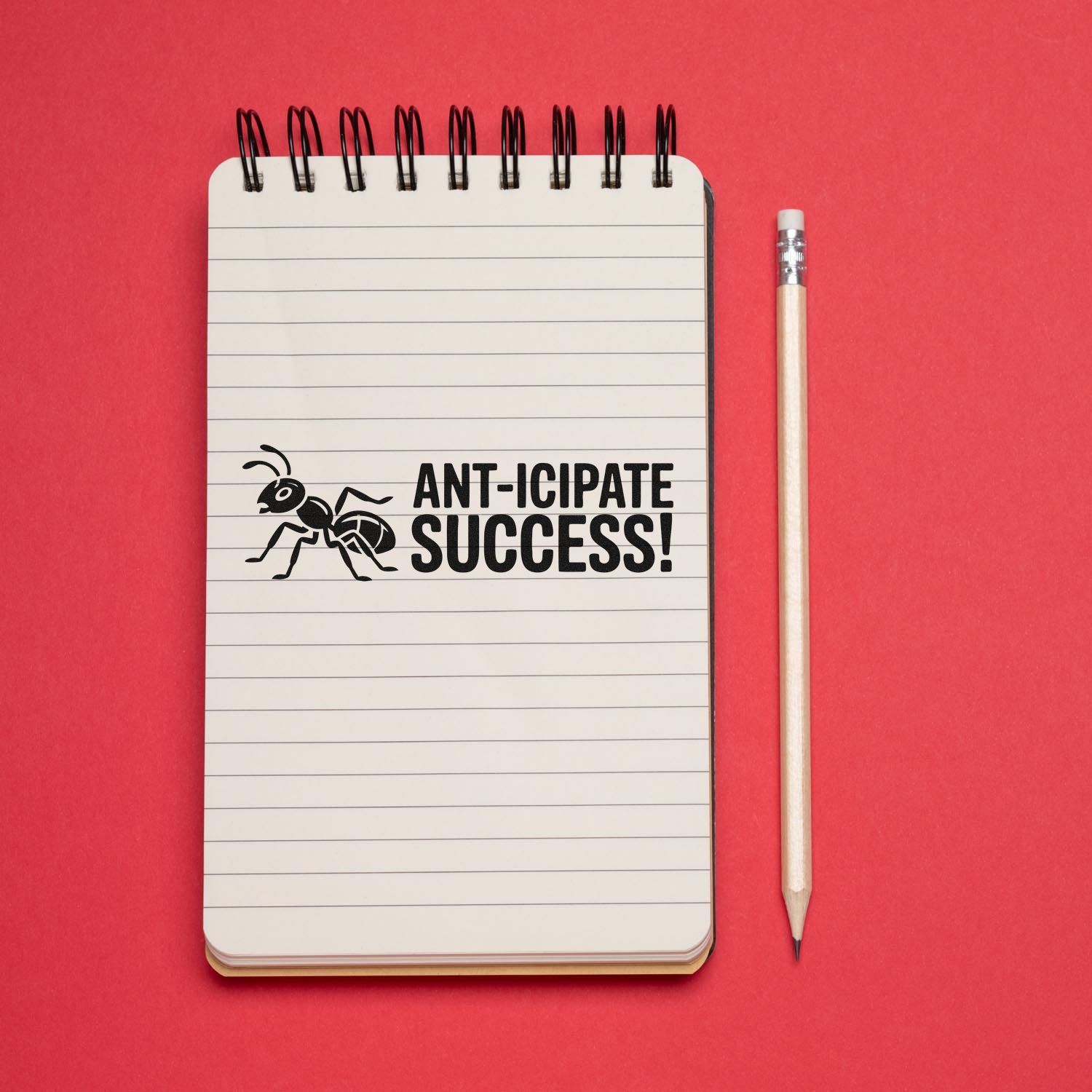 Motivational rubber stamp reading Ant-icipate Success! with ant icon on a lined notebook beside a pencil.