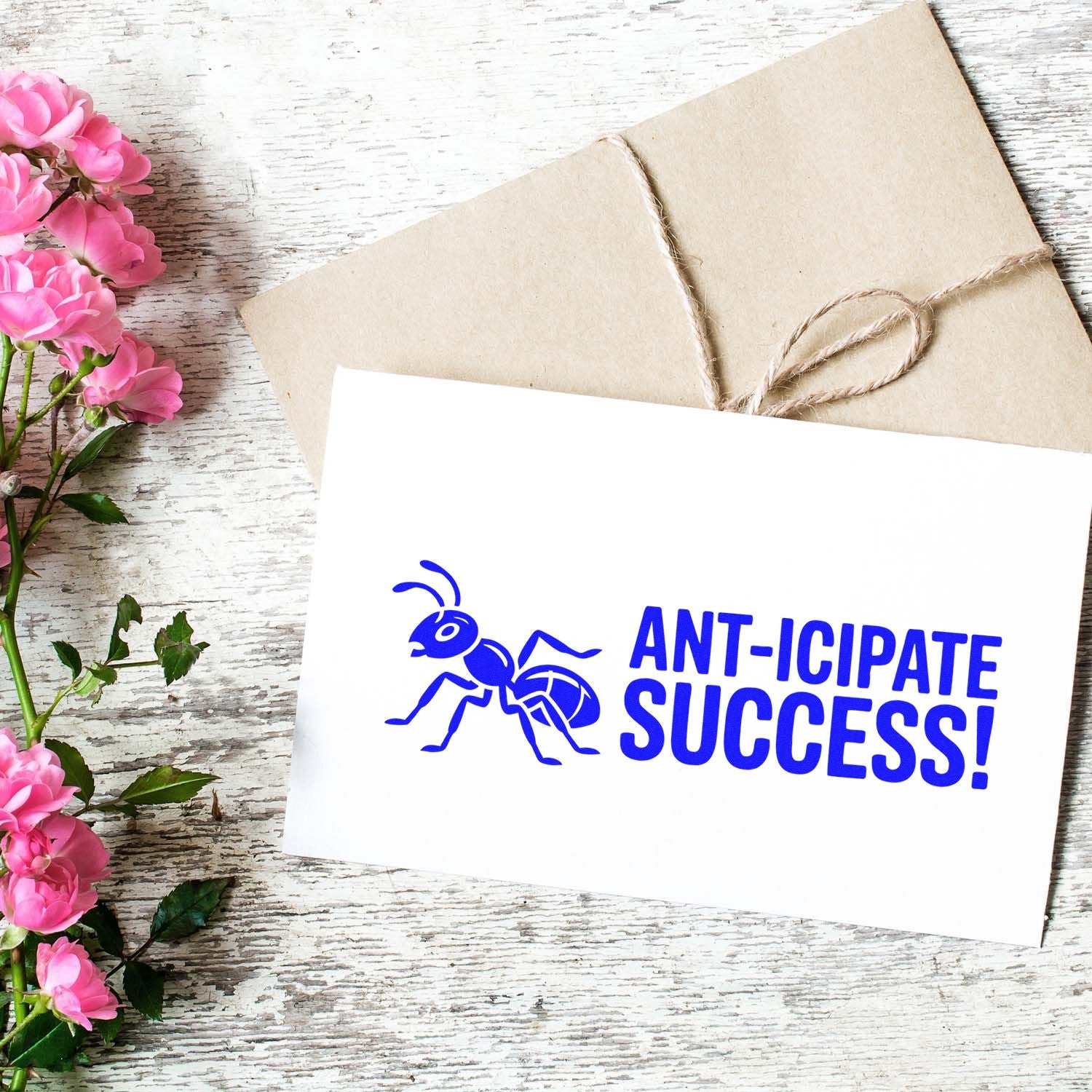 Ant rubber stamp design with Ant-icipate Success! on a card beside a wrapped envelope and pink flowers.