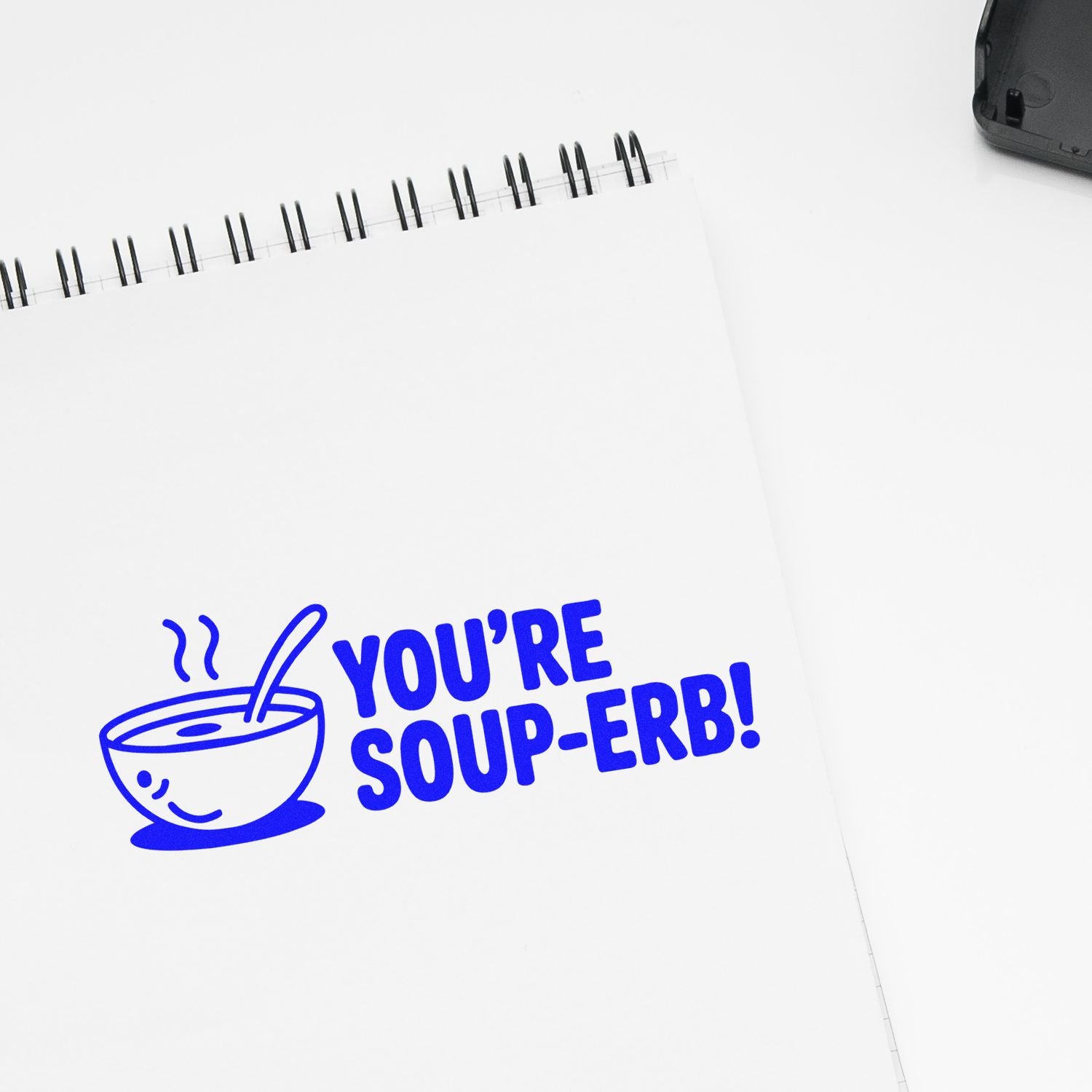 Blue rubber stamp on notebook reading Youre Soup-erb! with a smiling soup bowl, ideal for food-themed stationery and gifts.