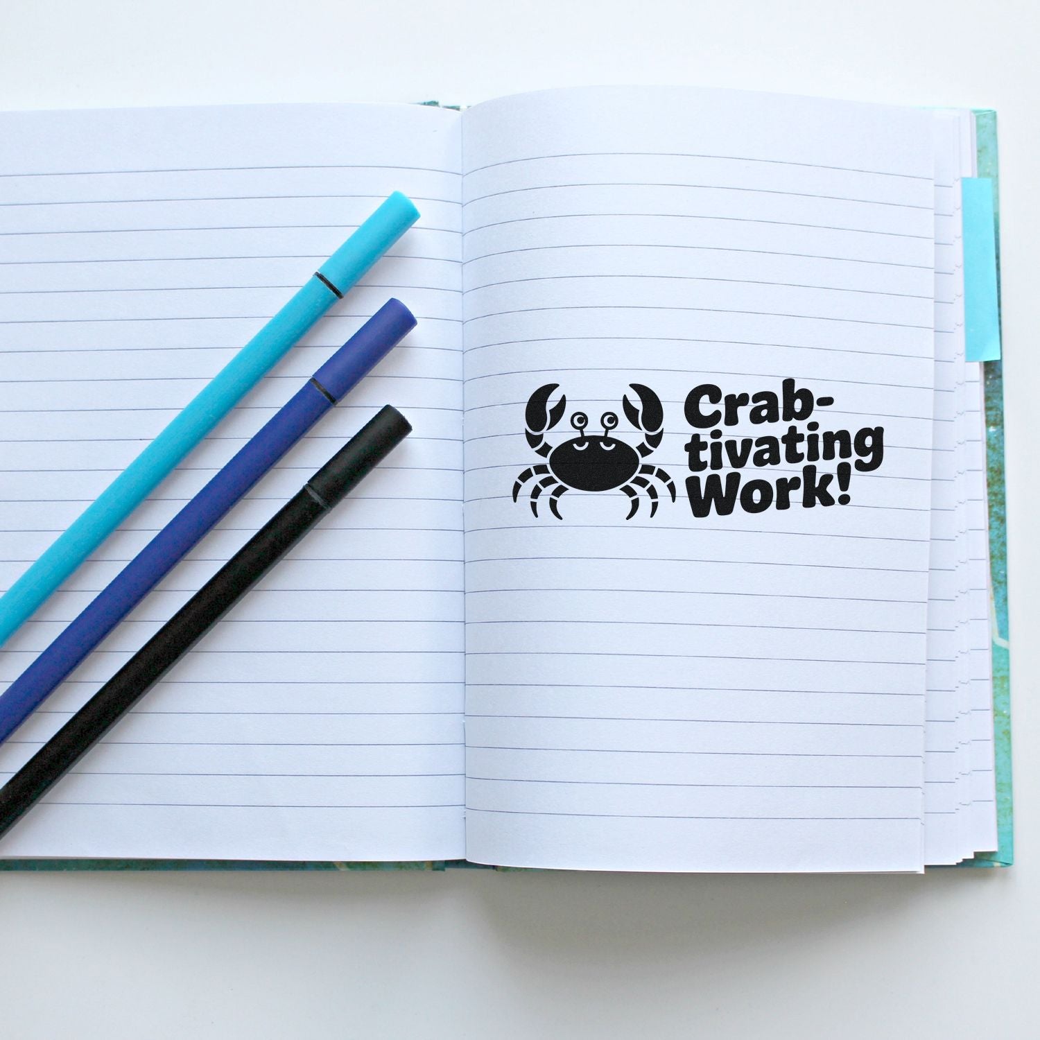 Crab-themed rubber stamp impression reading Crab-tivating Work! in an open notebook with pens, ideal for teacher or office use.