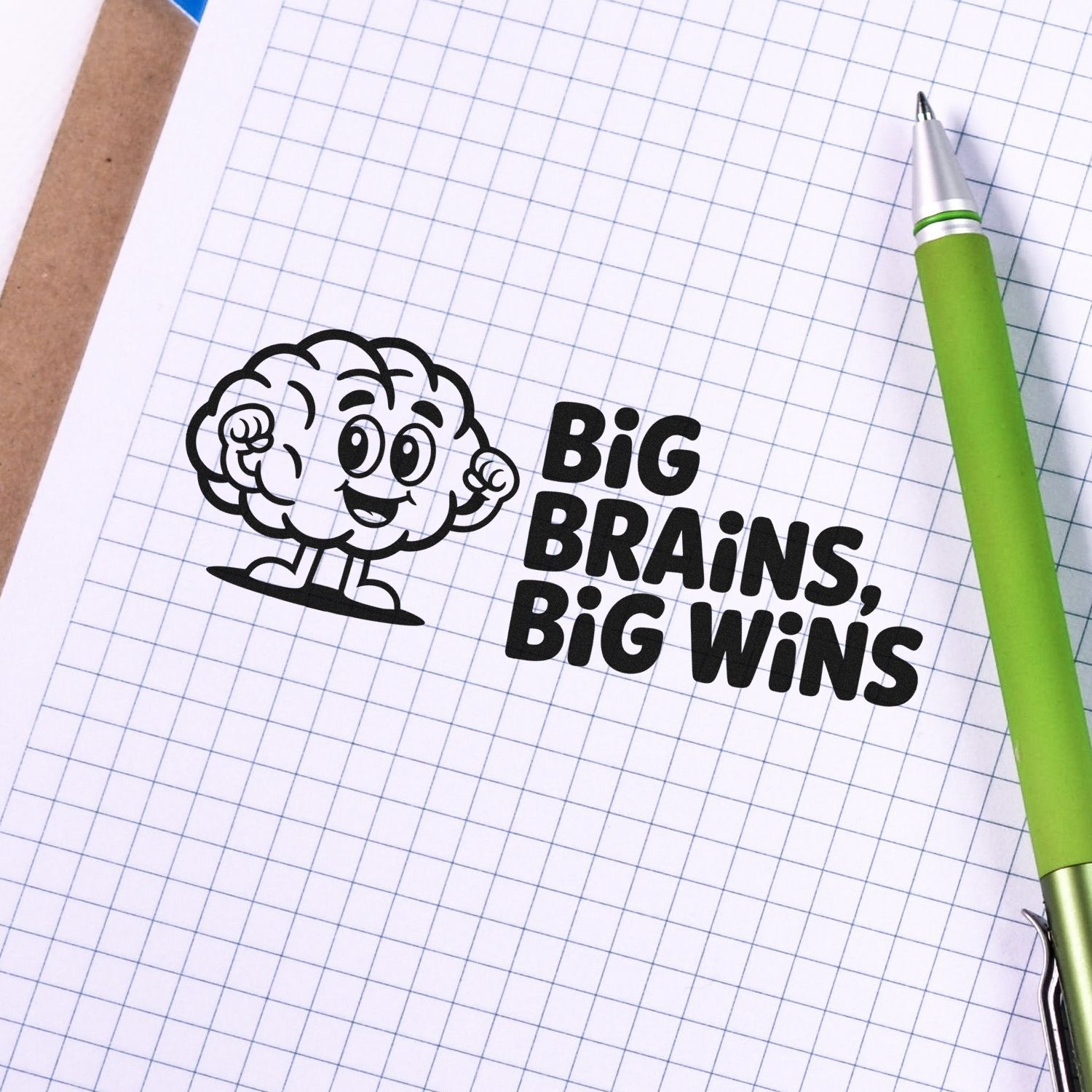 Custom rubber stamp reading Big Brains, Big Wins with cartoon brain design on graph paper beside a green pen.