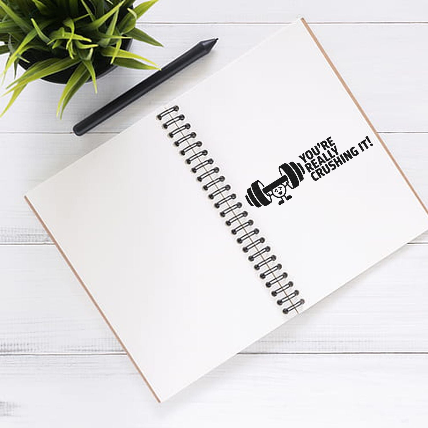 Motivational rubber stamp reading Youre really crushing it! with dumbbell icon on a spiral notebook.