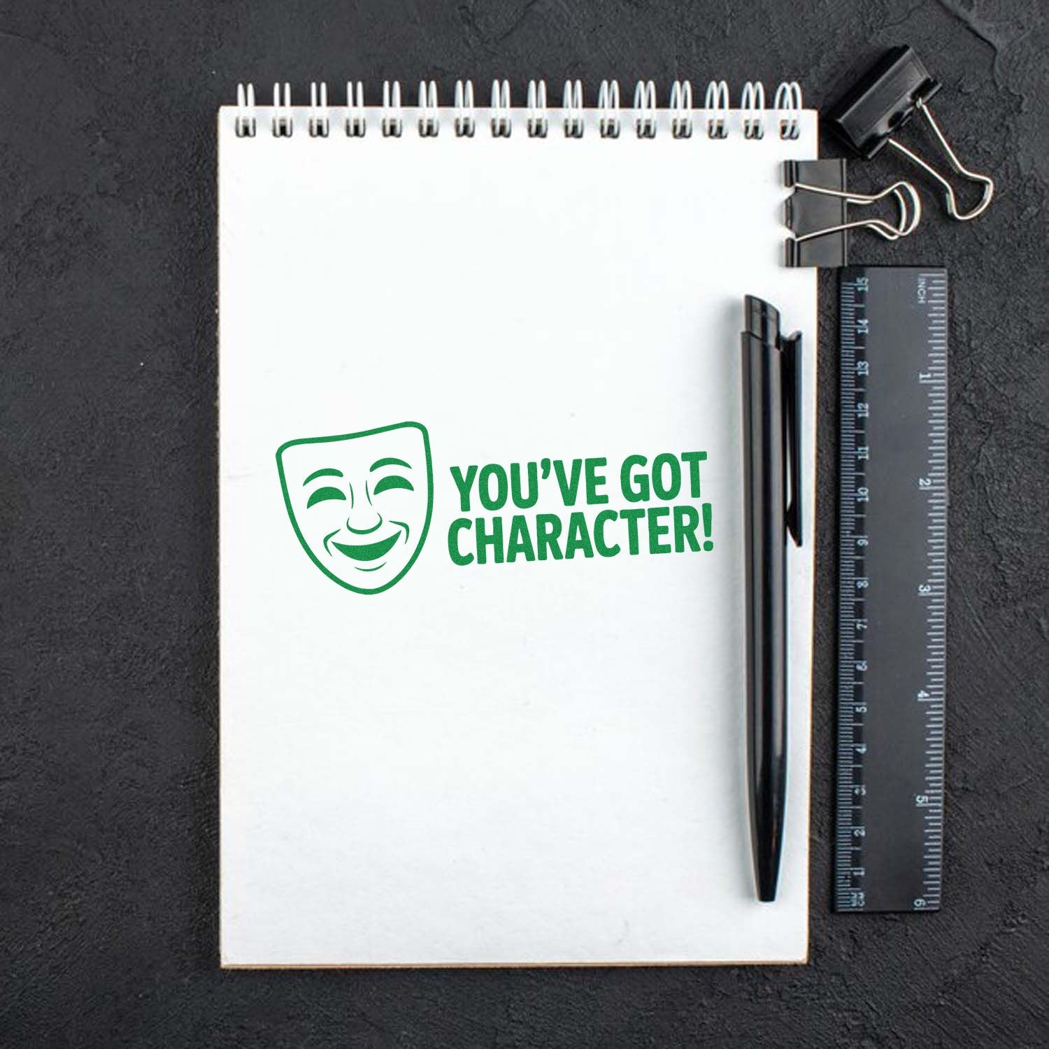 Green rubber stamp design with theater mask icon and Youve Got Character! message on a notebook, for stationery or craft use.