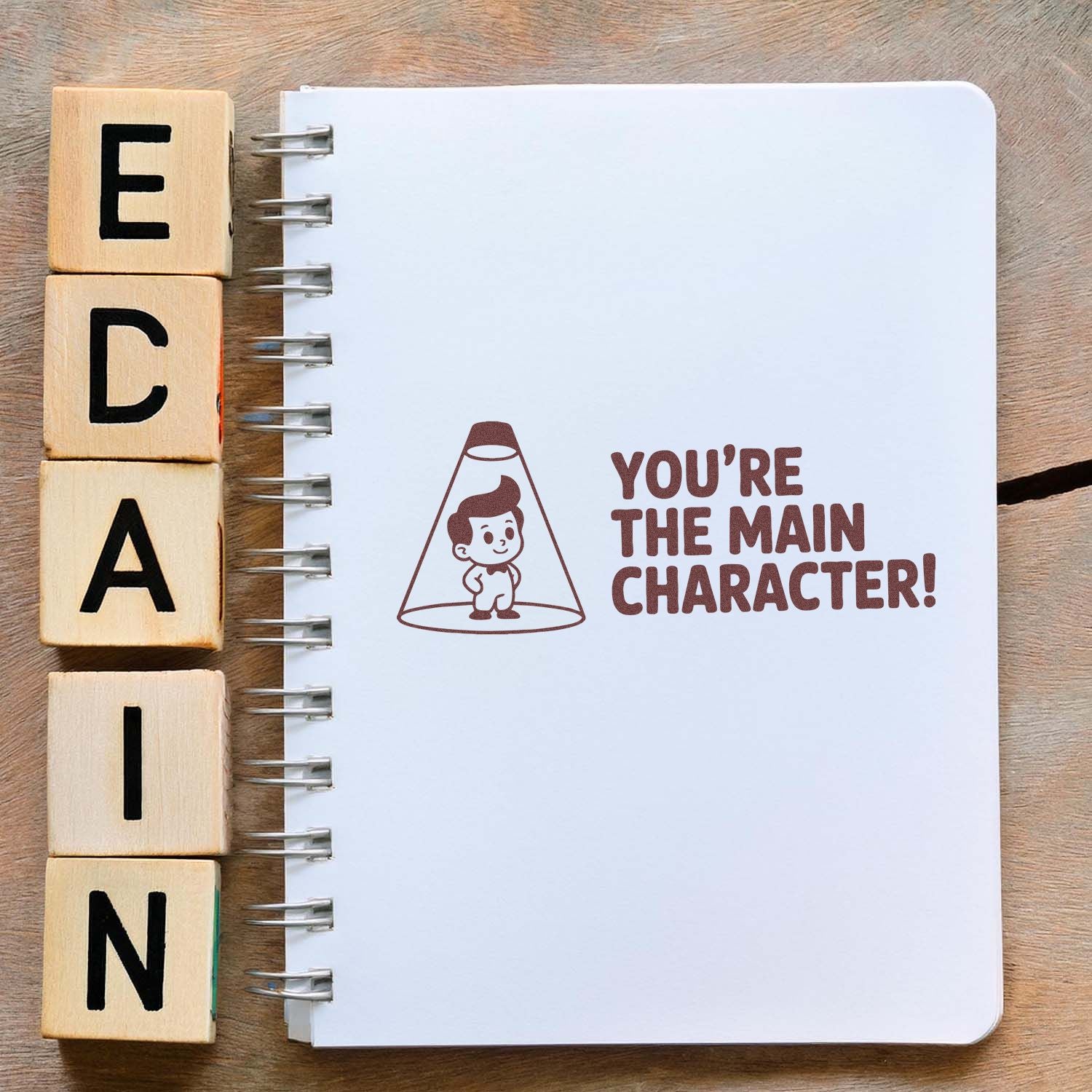 Custom rubber stamp mockup with wooden letter blocks and Youre the main character! design on a notebook page.