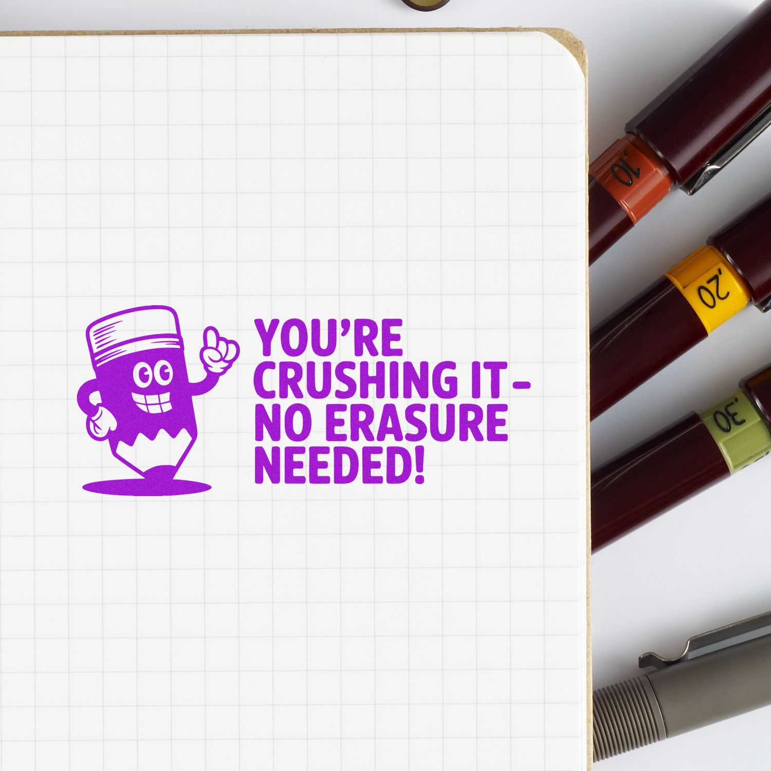 Motivational rubber stamp reading Youre crushing itno erasure needed! on notebook, with color-coded pen caps beside it.