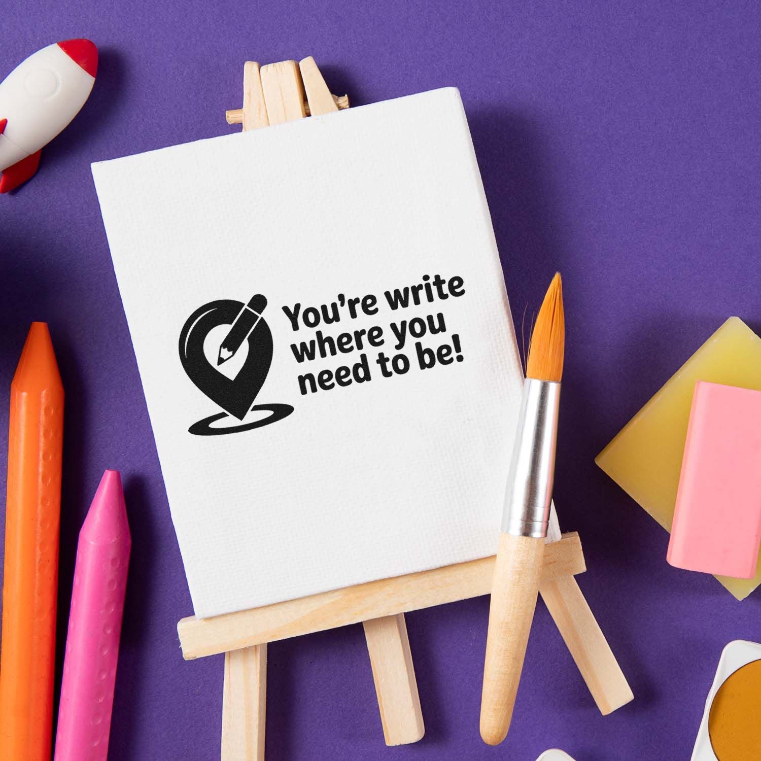 Custom rubber stamp design on mini easel with Youre write where you need to be! and art supplies on purple background.