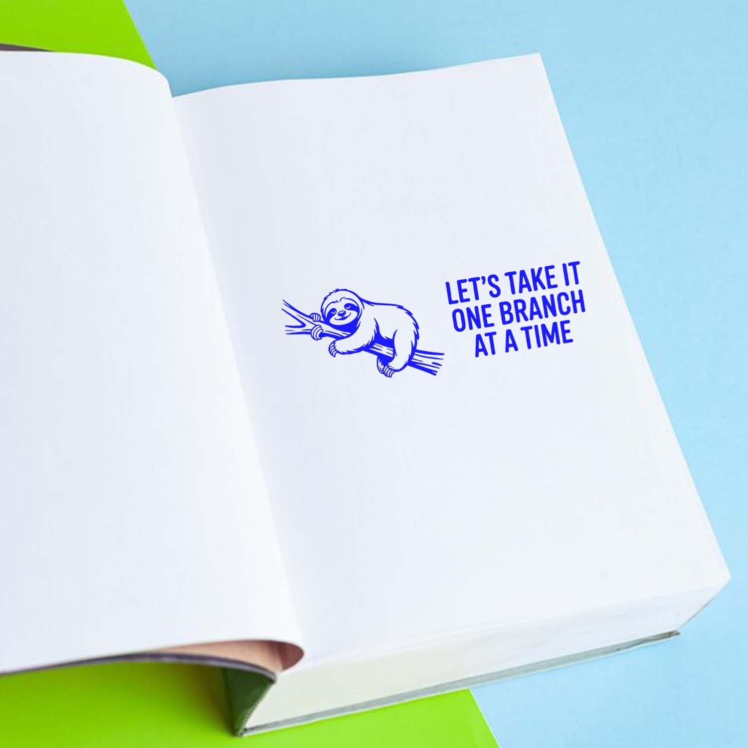 Sloth rubber stamp imprint with Lets Take It One Branch at a Time motivational message on open notebook.
