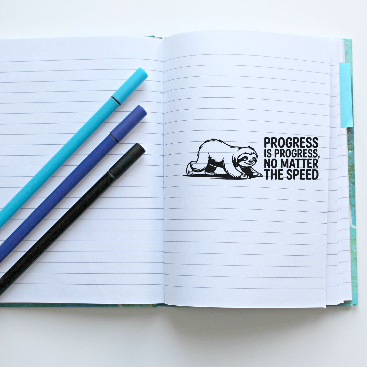 Sloth rubber stamp design on lined notebook page with pens, showing motivational quote: Progress is progress, no matter the speed.