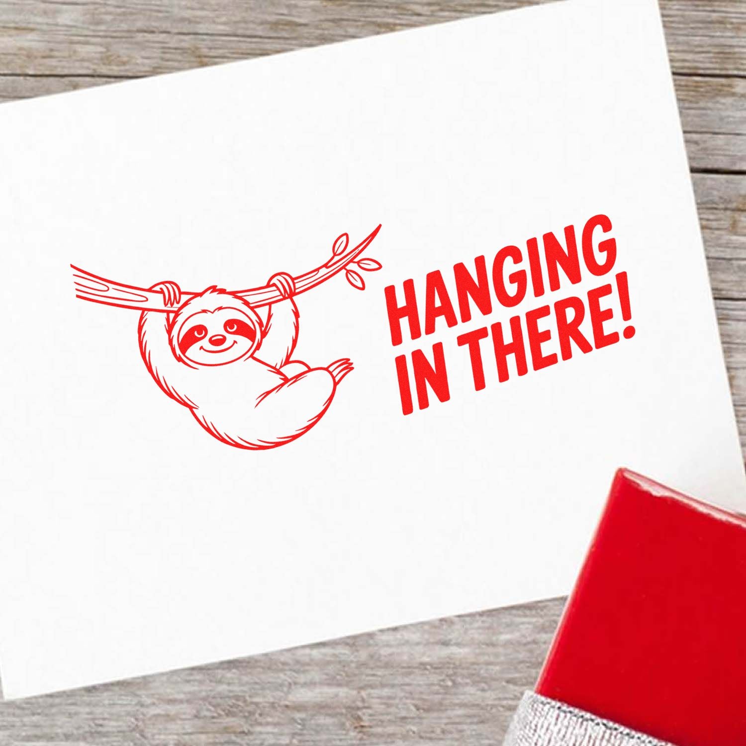 Motivational sloth rubber stamp with Hanging in There! text, shown as a red stamped impression on paper.