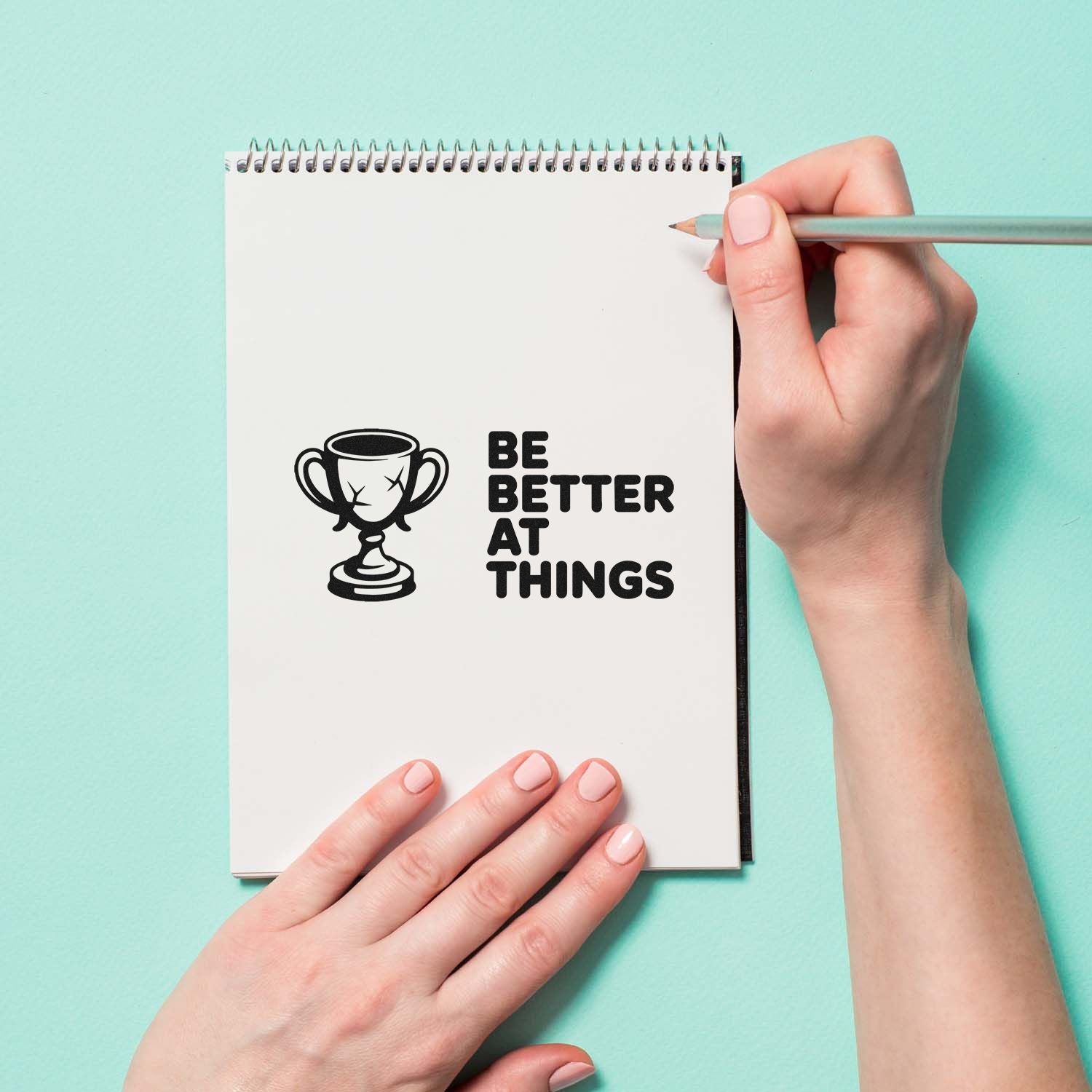 Motivational rubber stamp design with trophy icon and Be Better at Things quote on a notebook page.