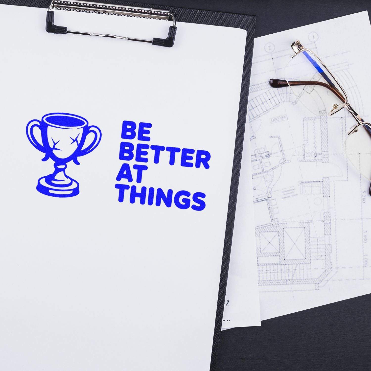 Custom rubber stamp with blue trophy and Be Better at Things slogan on clipboard, shown with blueprint and glasses.