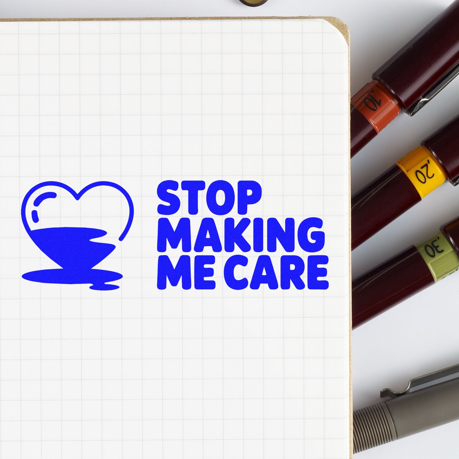 Rubber stamp design reading Stop Making Me Care with blue heart icon, shown on a notebook with ink stamp markers.