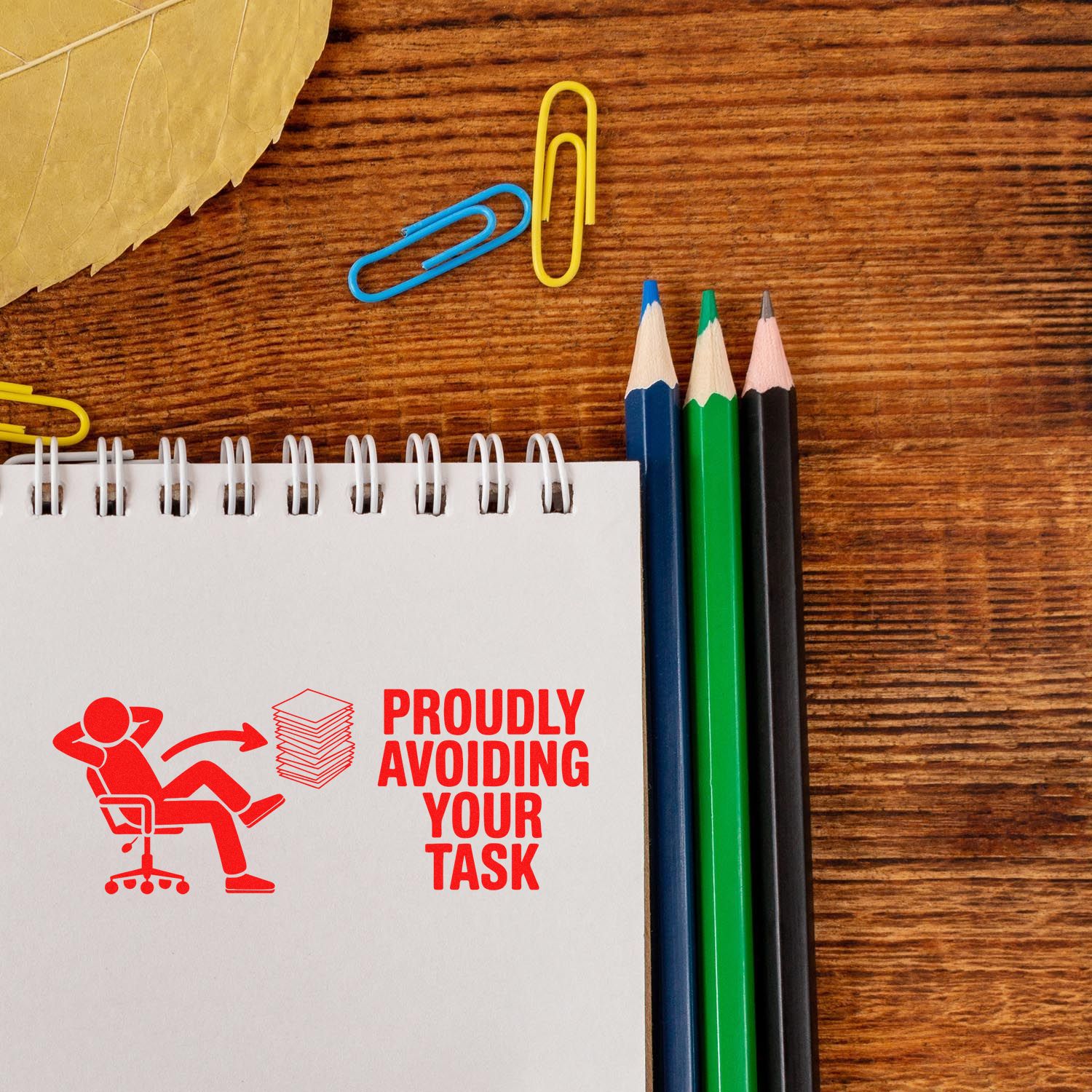 Funny custom rubber stamp reading Proudly Avoiding Your Task on a notebook with desk supplies.