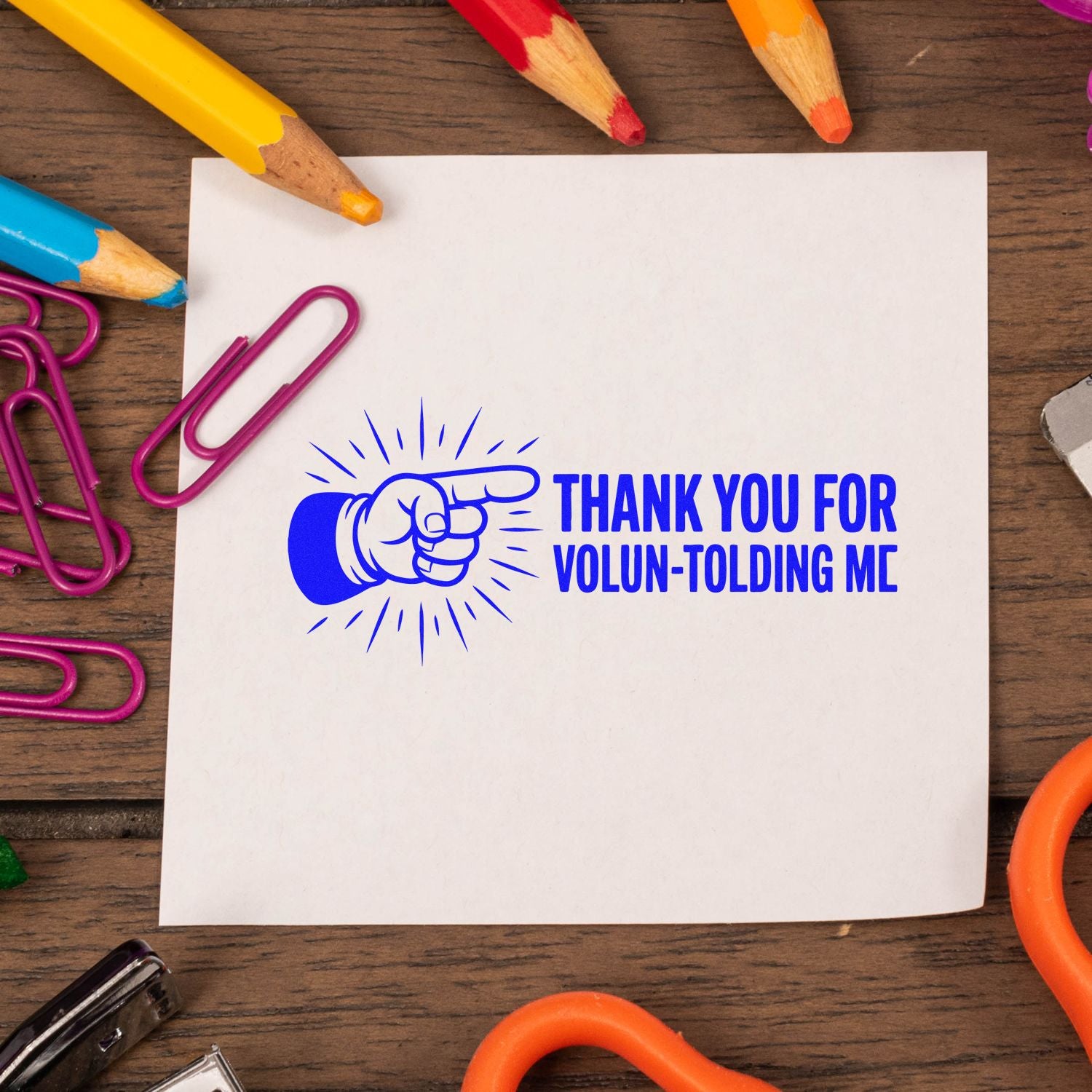 Volunteer thank-you rubber stamp with pointing hand and bold blue Thank You for Volun-tolding Me message on paper.