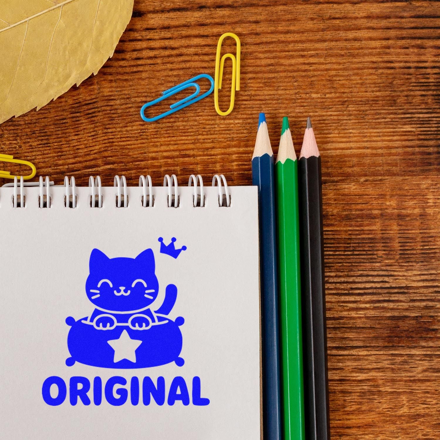 Custom rubber stamp design with blue cat illustration and Original on a notebook, surrounded by pencils and paper clips.