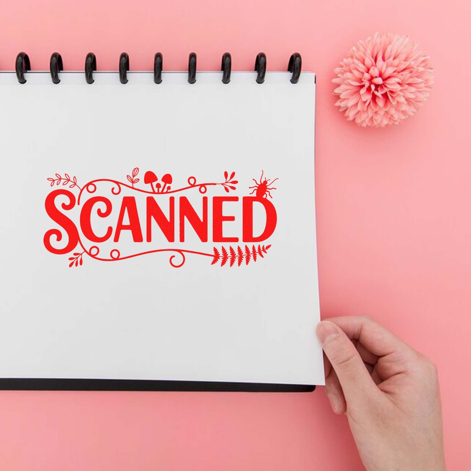 Red SCANNED decorative rubber stamp impression on notebook page, held in hand against a pink background.