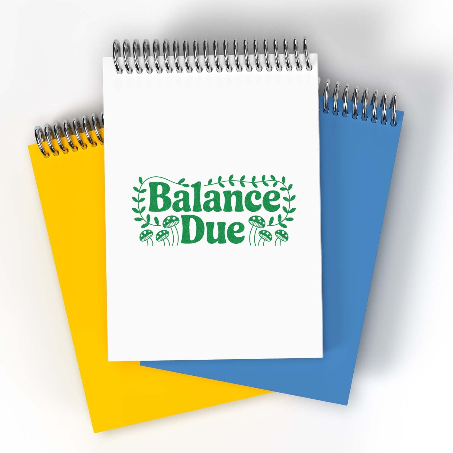 Balance Due rubber stamp design on a spiral notebook, with decorative leaves and mushrooms, shown on colorful notepads.