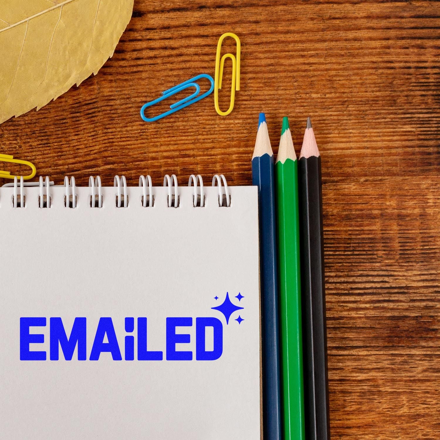 EMAILED rubber stamp on notepad beside pencils and paper clips on a wooden desk.